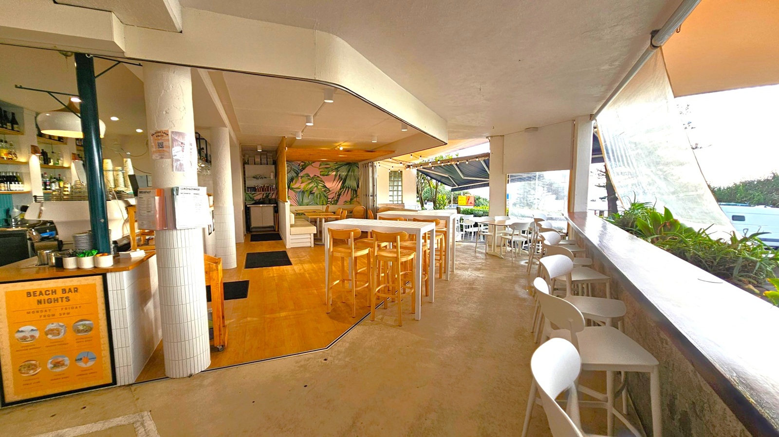 A spacious indoor-outdoor café with a mix of wooden and tiled flooring. On the left, there's a bar area with shelves of bottles and a coffee machine. A white column displays menus. In the centre, high wooden tables and stools are arranged, with a mural of tropical plants in the background. To the right, a long counter with plants overlooks outdoor seating with white chairs and tables. The ceiling is light-coloured with recessed lighting, and large windows provide natural light.