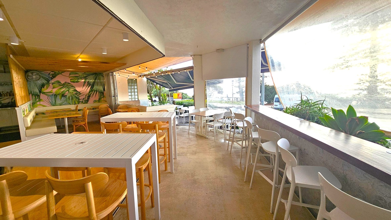 Open-air café with a tropical theme. On the left, a mural with large green leaves decorates the wall above a cushioned bench and wooden chairs. In the centre, white tables and wooden chairs are arranged on a concrete floor. The right side features a long counter with white stools and potted plants. Large windows with transparent blinds offer a view of greenery and a parked van outside. The ceiling is simple with recessed lighting.
