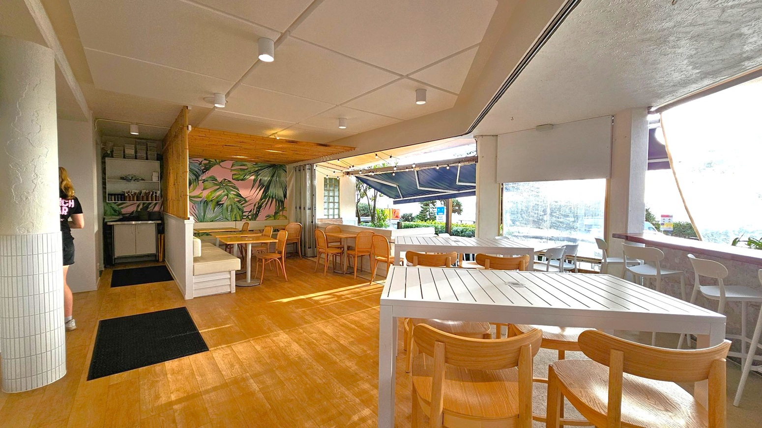 Interior of a café with wooden flooring and a white panelled ceiling. On the left, a person in a black shirt and shorts is walking past a column with white tiles. In the centre, there are tables with wooden chairs and a mural of green leaves on a pink background. The right side features large windows with views of greenery and the sea, partially covered by a blue awning. White tables and chairs are arranged near the windows, and string lights hang from the ceiling.