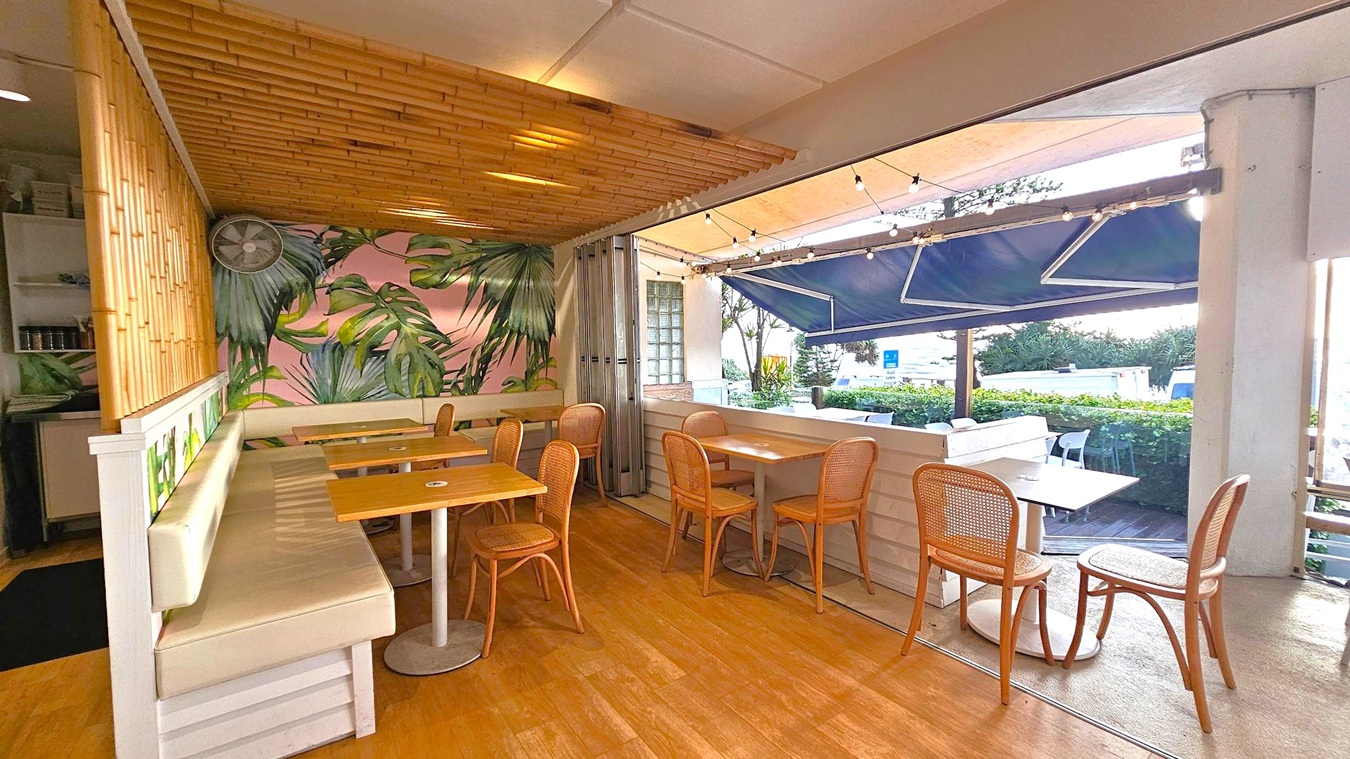 A cosy café interior with a tropical theme. On the left, there's a bamboo ceiling and wall, with a long cushioned bench and wooden tables. The wall features a large mural of green leaves. On the right, there are more wooden tables and chairs, with a view of the outside through large open windows. The exterior has a blue awning and string lights, with greenery visible beyond. The floor is wooden, and the ceiling is white.