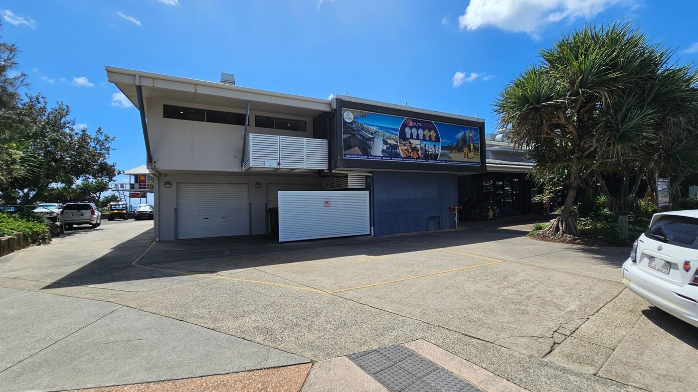 A two-storey building with a large sign on the upper right side, featuring images and text. The lower part has a closed roller door and a "No Entry" sign. To the left, there are parked cars and trees. On the right, a large tree with spiky leaves and a white car. The ground is a mix of textured concrete and green-painted sections. The sky is clear with a few clouds.