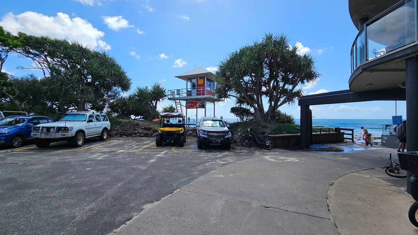 A coastal scene with a parking area in the foreground, featuring several vehicles including a white SUV, a yellow utility vehicle, and a black car. In the centre, a lifeguard tower stands elevated among trees. To the right, a curved building with glass balconies overlooks the ocean. A person is visible near the beach access, and a bicycle is parked nearby. The ocean is visible in the background.