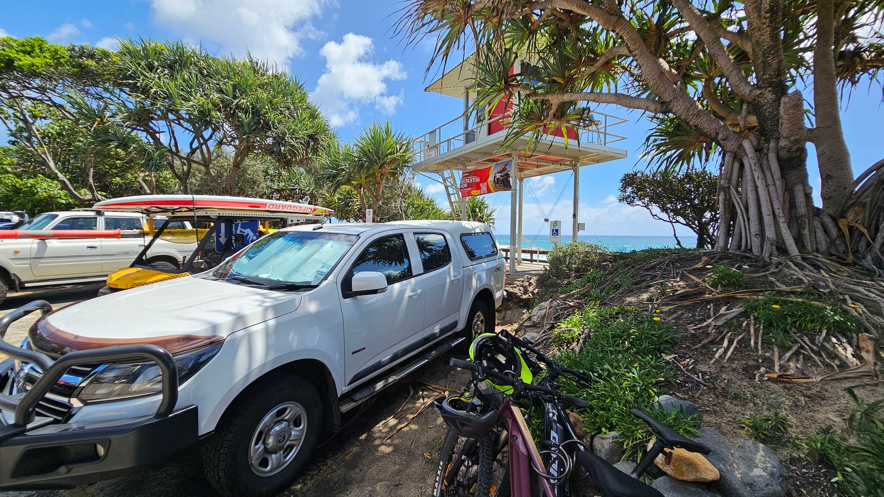 A white utility vehicle is parked on the left, with a lifeguard surfboard mounted on top. Next to it, a yellow lifeguard buggy is visible. On the right, a lifeguard tower stands elevated, with a red and white design. In the foreground, there are bicycles parked near a large tree with exposed roots. The background features lush green trees and a glimpse of the ocean.