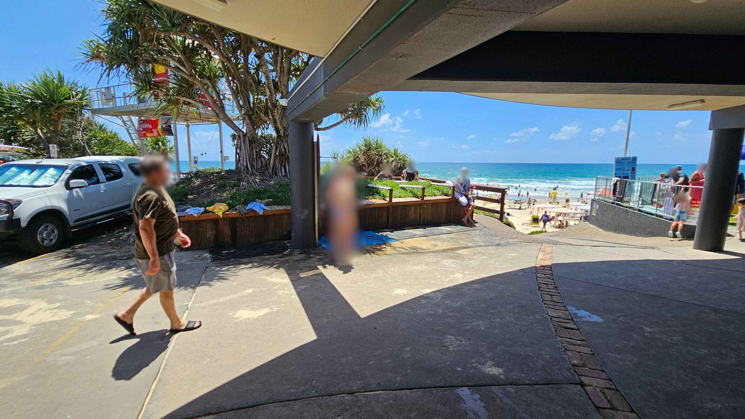A beach scene with a man walking on a paved area in the foreground. To the left, a white vehicle is parked near a lifeguard tower surrounded by trees. In the centre, a man in swimwear stands near a wooden fence with towels draped over it. The background features a sandy beach with people enjoying the ocean. A shaded structure covers part of the area, casting shadows on the ground.