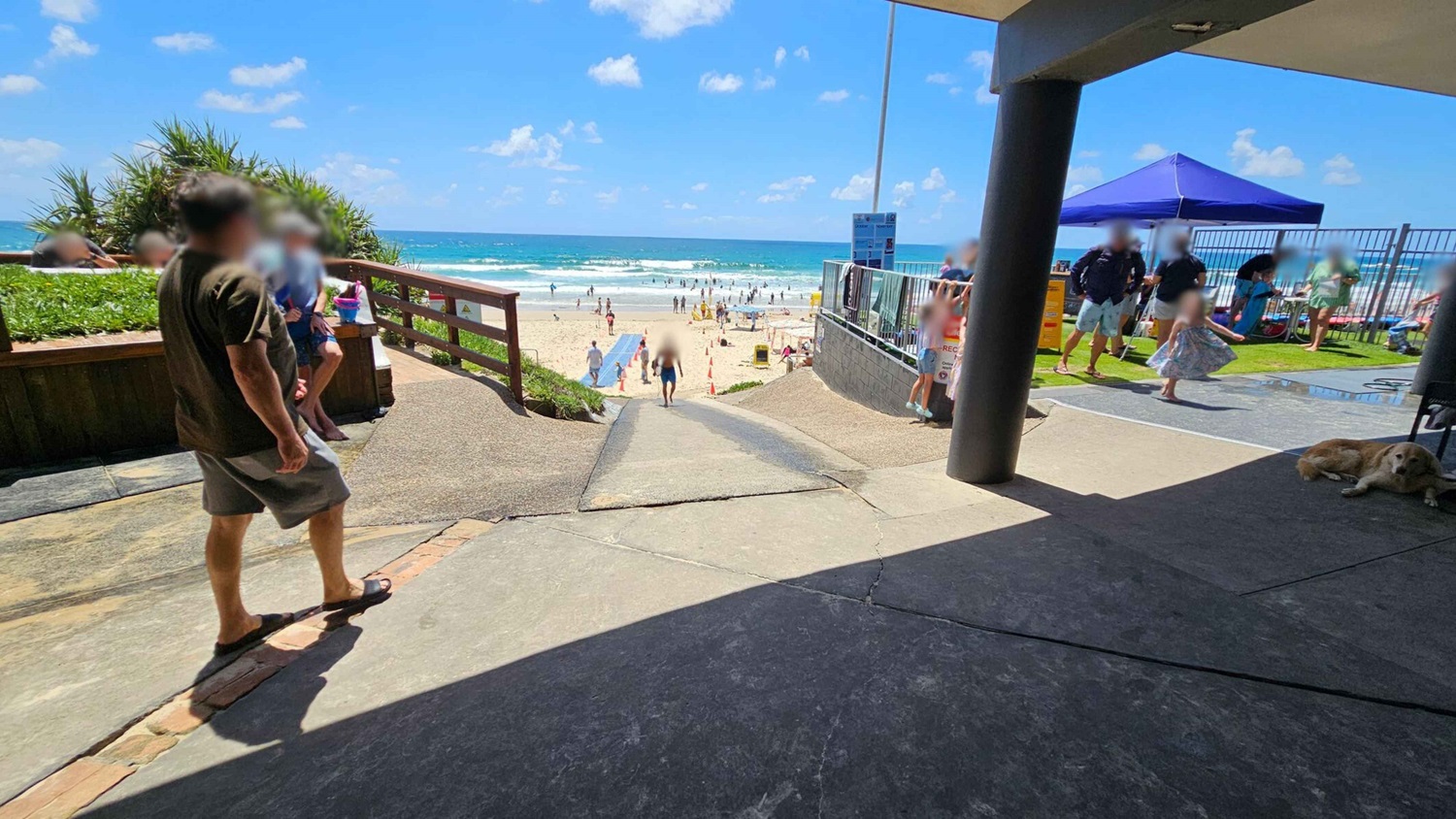 A beach scene with people enjoying the sunny day. On the left, a man walks past a wooden railing with greenery. In the centre, a path leads to the sandy beach where people are swimming and sunbathing. On the right, a blue tent is set up on a grassy area with people gathered around. A child in a dress is running, and a dog is lying on the ground near a column. The ocean is visible in the background with gentle waves.