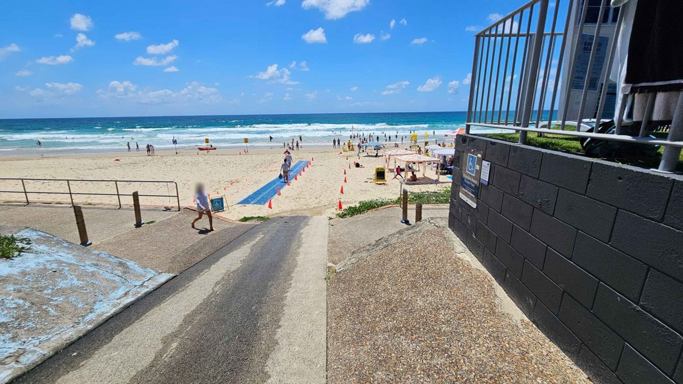 A beach scene with a pathway leading to the sand. On the right, there's a black brick wall with a railing above it. A sign indicating accessibility is visible. On the left, a sloped path with a railing runs parallel to the beach. People are scattered across the sand, with some near the water. A blue mat extends towards the sea, and orange cones mark areas on the sand. The ocean is in the background with gentle waves.