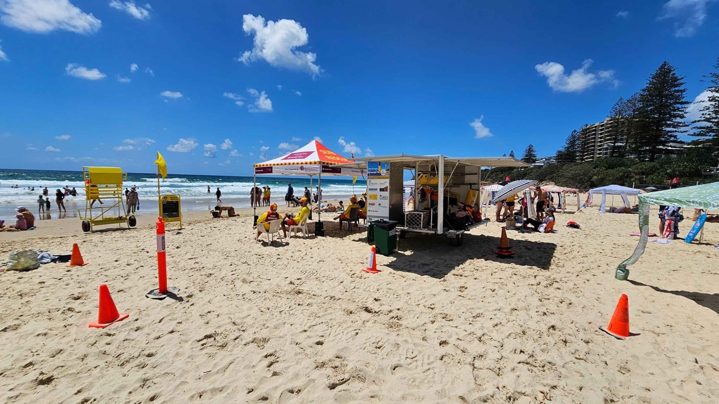 A sandy beach with a lifeguard station in the centre, featuring a red and yellow tent and a trailer with equipment. Lifeguards in yellow and red uniforms sit under the tent. To the left, people are swimming in the sea, and a yellow lifeguard chair is visible. On the right, beachgoers are under small canopies. Orange cones are scattered on the sand. In the background, there are trees and a few tall buildings.
