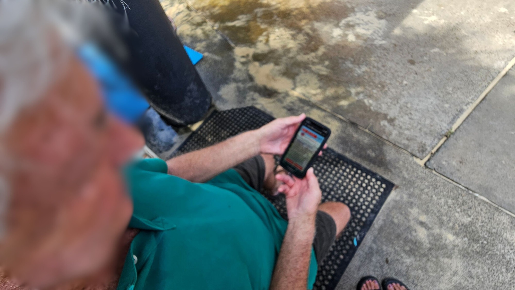 A person with grey hair is sitting on a black mat, holding a smartphone. They are wearing a green shirt and shorts. The ground is made of concrete with patches of discolouration. A pair of feet in sandals is visible at the bottom right. A blue object and a dark vertical structure are on the left side.