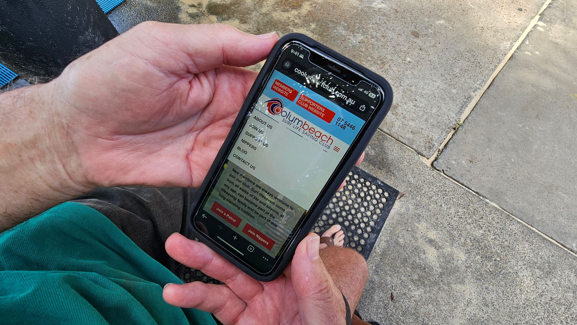 A person is holding a smartphone displaying the Coolum Beach Surf Life Saving Club website. The phone is in a black case, and the screen shows menu options like "About Us" and "Join Us". The person is wearing a green and black garment. In the background, there is a concrete surface with patches of green and a small black mat with circular patterns. A pair of feet in sandals is partially visible.