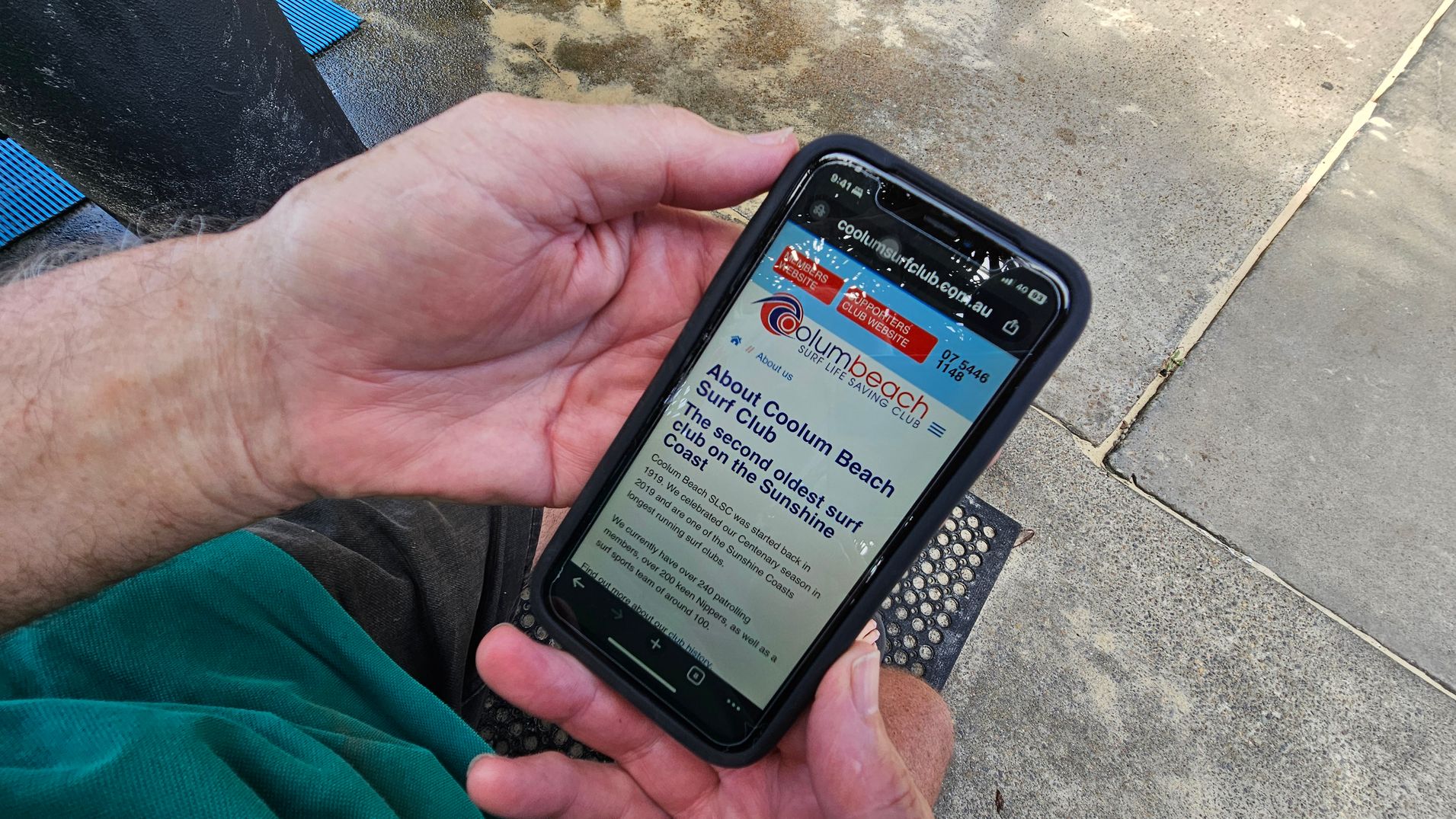 A person is holding a smartphone displaying a webpage about the Coolum Beach Surf Club. The phone is in a black case, and the person's hands are visible. They are wearing a green shirt and grey shorts. The background shows a concrete surface with patches of green moss or algae, and a small black mat with circular patterns is partially visible.
