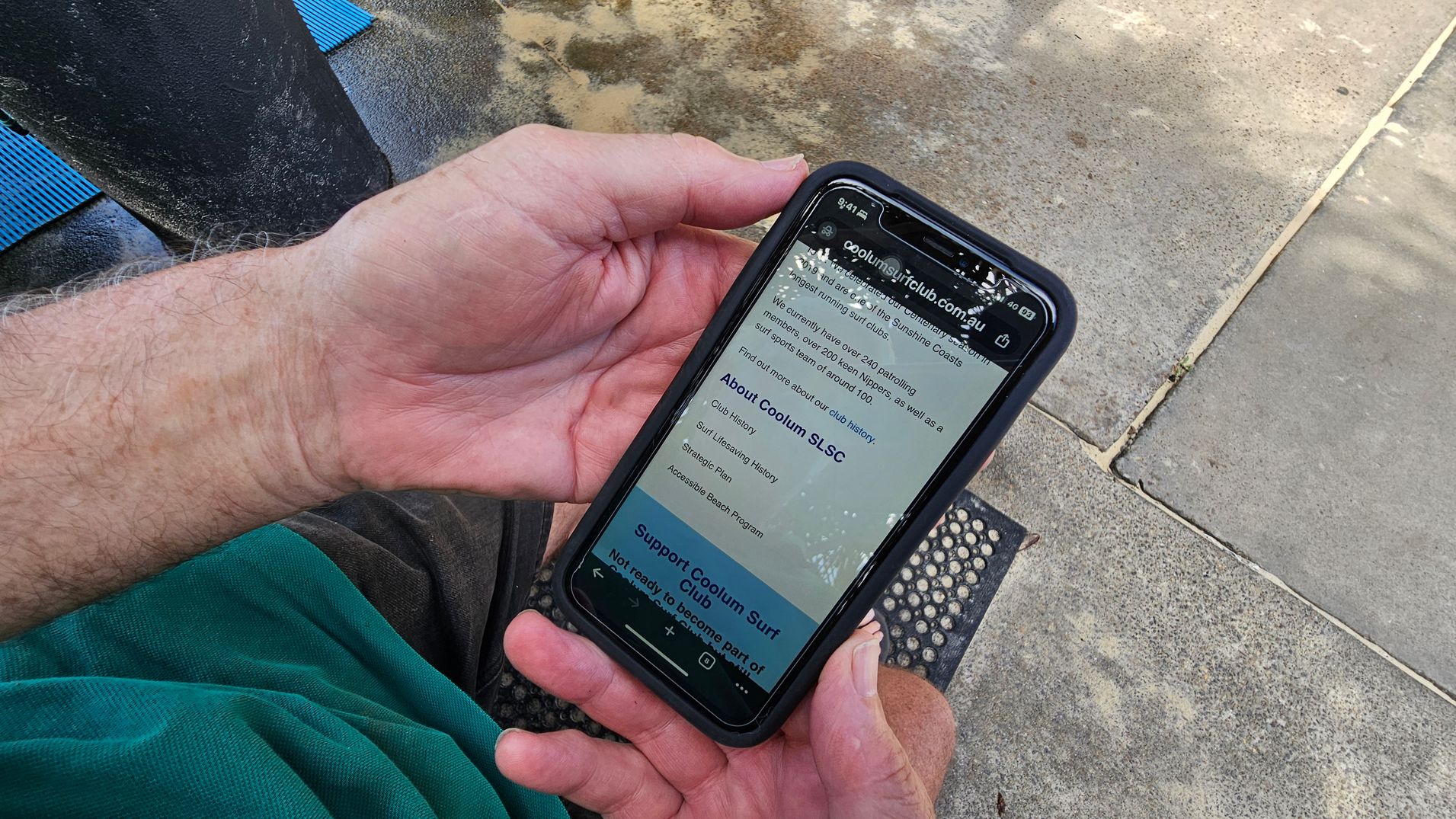 A person is holding a smartphone displaying a website about the Coolum Surf Club. The phone is in a black case, and the person is wearing a green shirt. The background shows a concrete surface with patches of moss or lichen, and a blue mat is partially visible on the left.