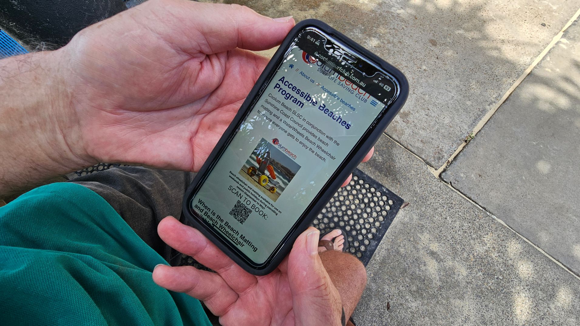A person is holding a smartphone displaying a webpage about an "Accessible Beaches Program." The phone is held with both hands, and the screen shows text and an image of a beach wheelchair. The person is wearing a green shirt and grey shorts, sitting on a concrete surface with a textured mat nearby. The ground has patches of moss or lichen, and there are shadows from nearby foliage.