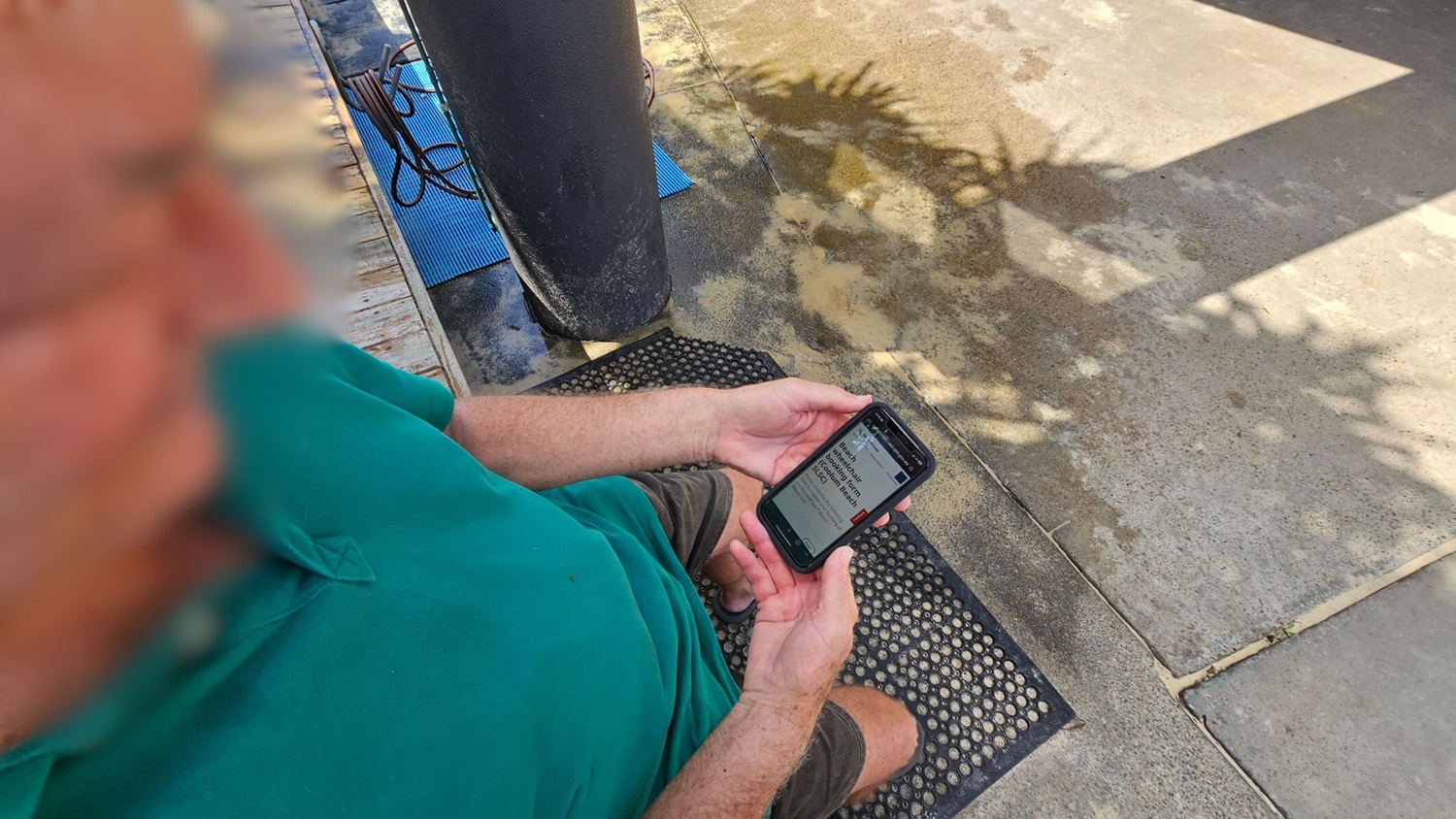 A person in a green shirt is sitting on a wooden deck, holding a smartphone. The screen displays text. To the right, there's a textured mat on the ground, and a large black column. The ground is a mix of concrete and wooden planks, with shadows of leaves creating patterns. A blue mat and coiled hose are visible in the background.
