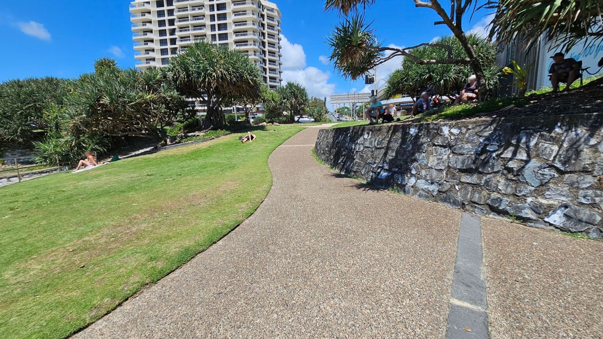 A curved pathway runs through a grassy area. On the left, people are sunbathing on the grass near dense trees. On the right, a stone wall borders the path, with people sitting above it under more trees. In the background, a tall building rises against a clear blue sky.