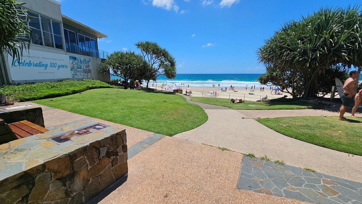 A beach scene with a grassy area in the foreground, featuring a stone and brick bench on the left. A path leads towards the sandy beach in the background, where people are enjoying the ocean. On the left, a building with large windows displays a sign celebrating 100 years. Trees with dense foliage are on both sides, providing shade. The ocean is visible in the distance, with waves and beachgoers.