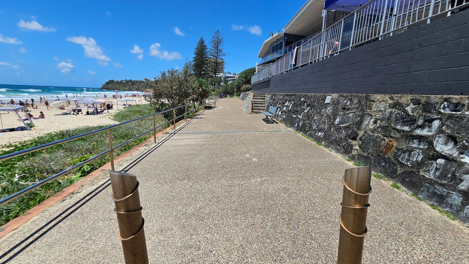 A coastal walkway with a textured path runs alongside a beach on the left, where people are sunbathing and swimming. A metal railing separates the path from the sandy beach. On the right, a stone wall supports a building with a balcony and railings. A bench is positioned along the path, and there are trees and shrubs in the background.