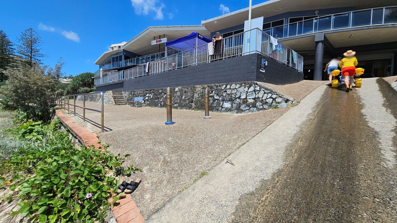 A building with a large balcony and glass railings is on the right. Two people are pushing a wheelchair with large yellow wheels up a ramp. On the left, there is a path with a metal railing and a small garden with green plants and a pair of sandals. The ground is a mix of concrete and gravel, and there is a stone wall supporting the building. A blue sky is visible above.