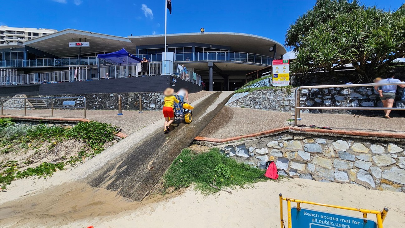 A sandy beach in the foreground leads to a ramp with a textured surface. Two people are pushing a beach wheelchair up the ramp. On the right, a stone wall with a sign and a tree. In the background, a modern building with a large overhang and glass windows. A blue canopy is set up on the left side of the building, and a few people are visible on the balcony. A yellow sign in the foreground reads "Beach access mat for all public users."