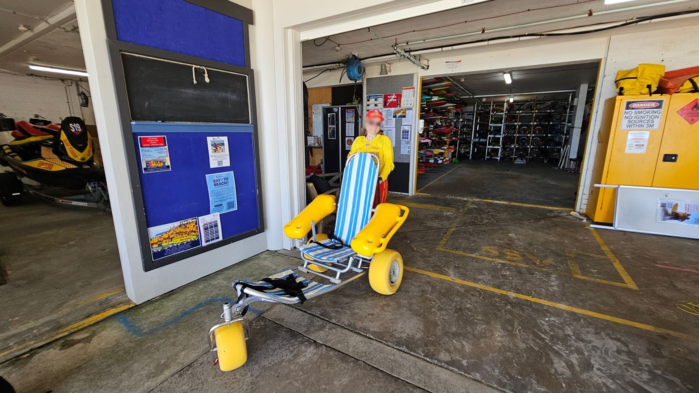 On the left, a blue noticeboard with posters is mounted on a white wall next to a parked yellow-and-black jet ski inside a garage. On the right, a yellow storage cabinet with a warning sign is positioned against the wall. In the middle, a striped blue-and-white beach wheelchair with yellow wheels is on a concrete floor, with a person in a yellow shirt standing behind it. The background includes a storage area with kayaks and equipment on racks.