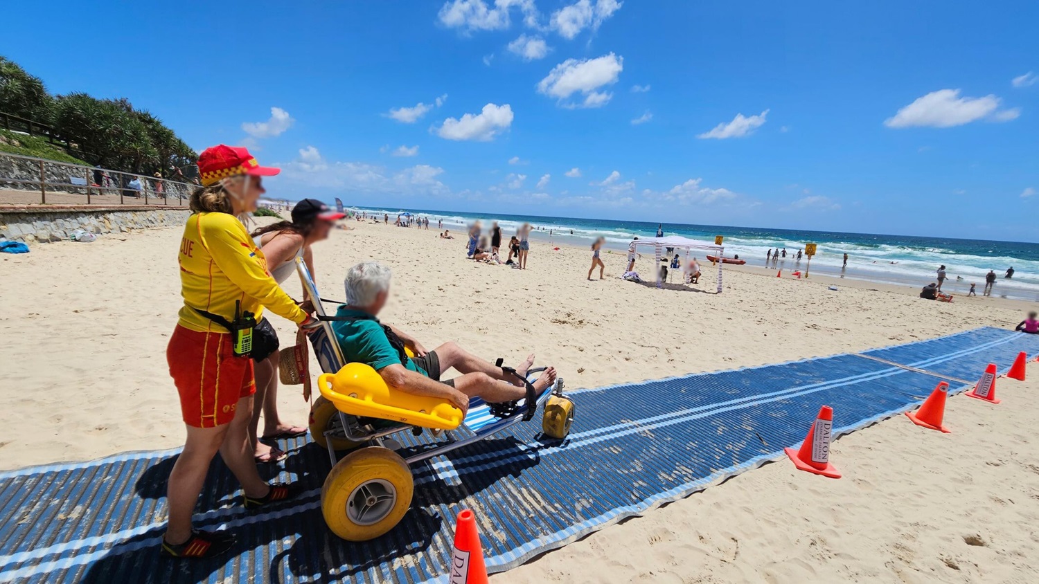 A man is seated in a beach wheelchair with large yellow wheels on a blue mat laid over the sand. Two women stand behind him, one wearing a lifeguard uniform. Orange cones mark the path. In the background, a stone wall with a railing separates the beach from a grassy area. A sign with beach safety information is visible, and there are people and beachgoers in the distance near the water.