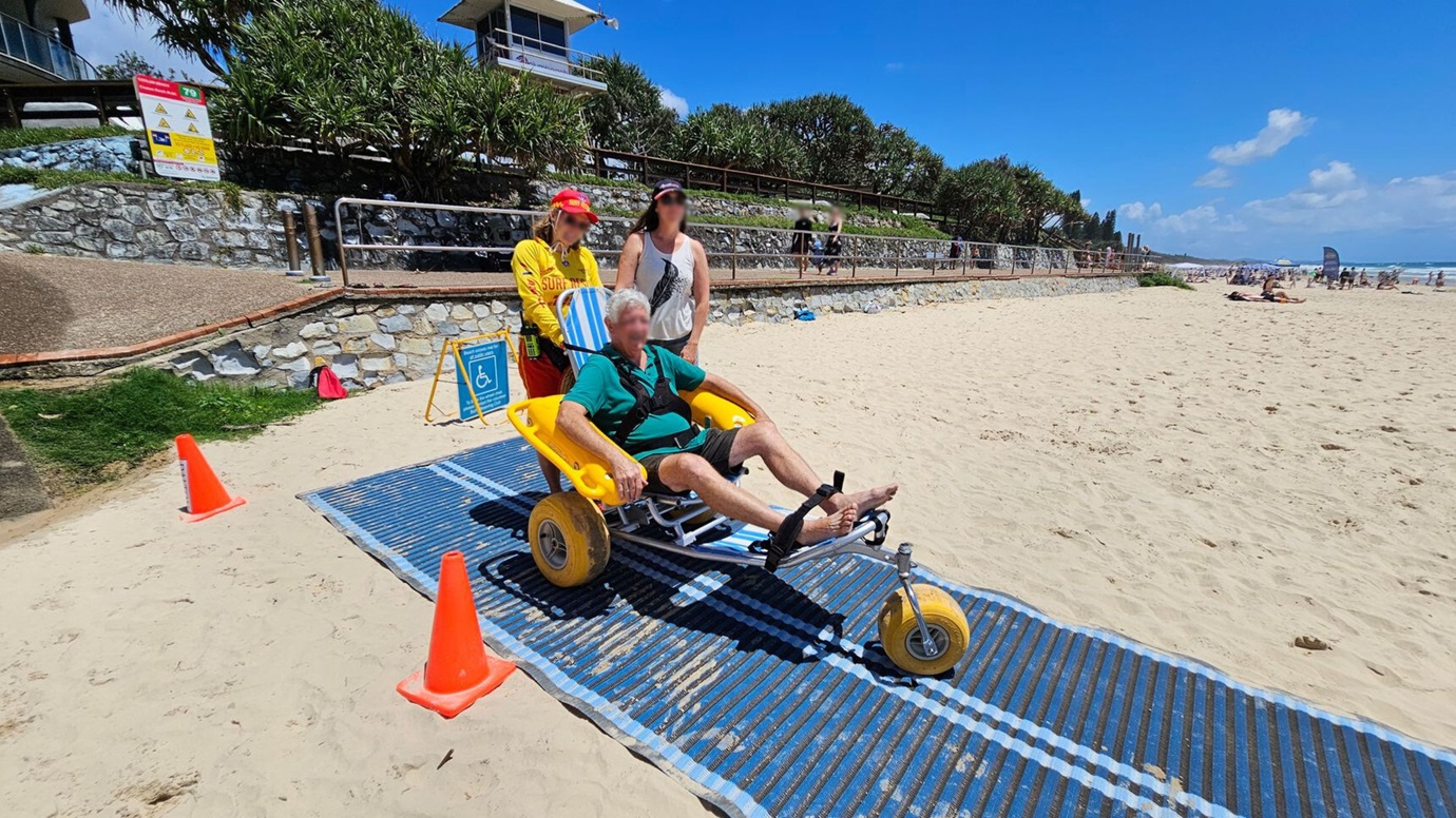 A beach scene with a sandy foreground and a blue mat pathway leading towards the ocean. On the left, a person in a yellow and red lifeguard uniform assists a man in a beach wheelchair with large yellow wheels. Another person stands nearby. The background shows people enjoying the beach and the sea, with some sitting under a canopy. Orange cones line the pathway. A stone wall and greenery are visible on the far left.