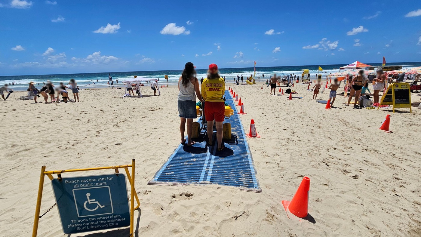 A sandy beach with a blue access mat in the centre, leading towards the ocean. Two people, one in a "Surf Rescue" shirt, are pushing a beach wheelchair along the mat. Orange cones line the path. On the left, people are gathered near the water, some standing and others sitting. On the right, a group of people are near a yellow sign and a tent.