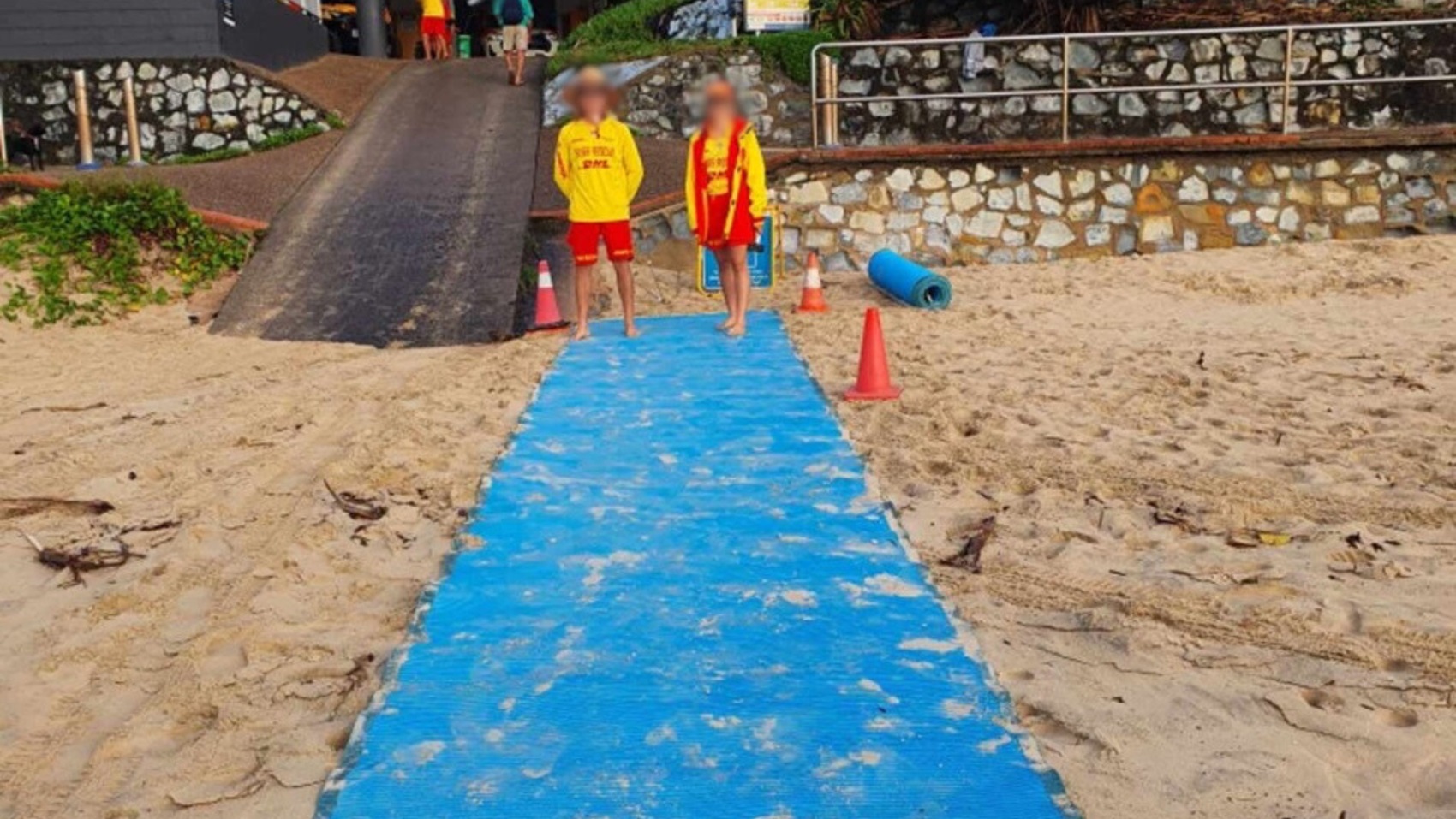 A blue mat stretches along the sandy ground in the foreground, bordered by sand on both sides. Two people in yellow and red uniforms stand in the middle, with small orange traffic cones nearby. On the left, a sloped black ramp leads up to a dark structure. On the right, a stone wall with a metal railing runs parallel to the ramp, with a blue rolled mat resting nearby. Greenery is visible at the edges of the ramp and wall.