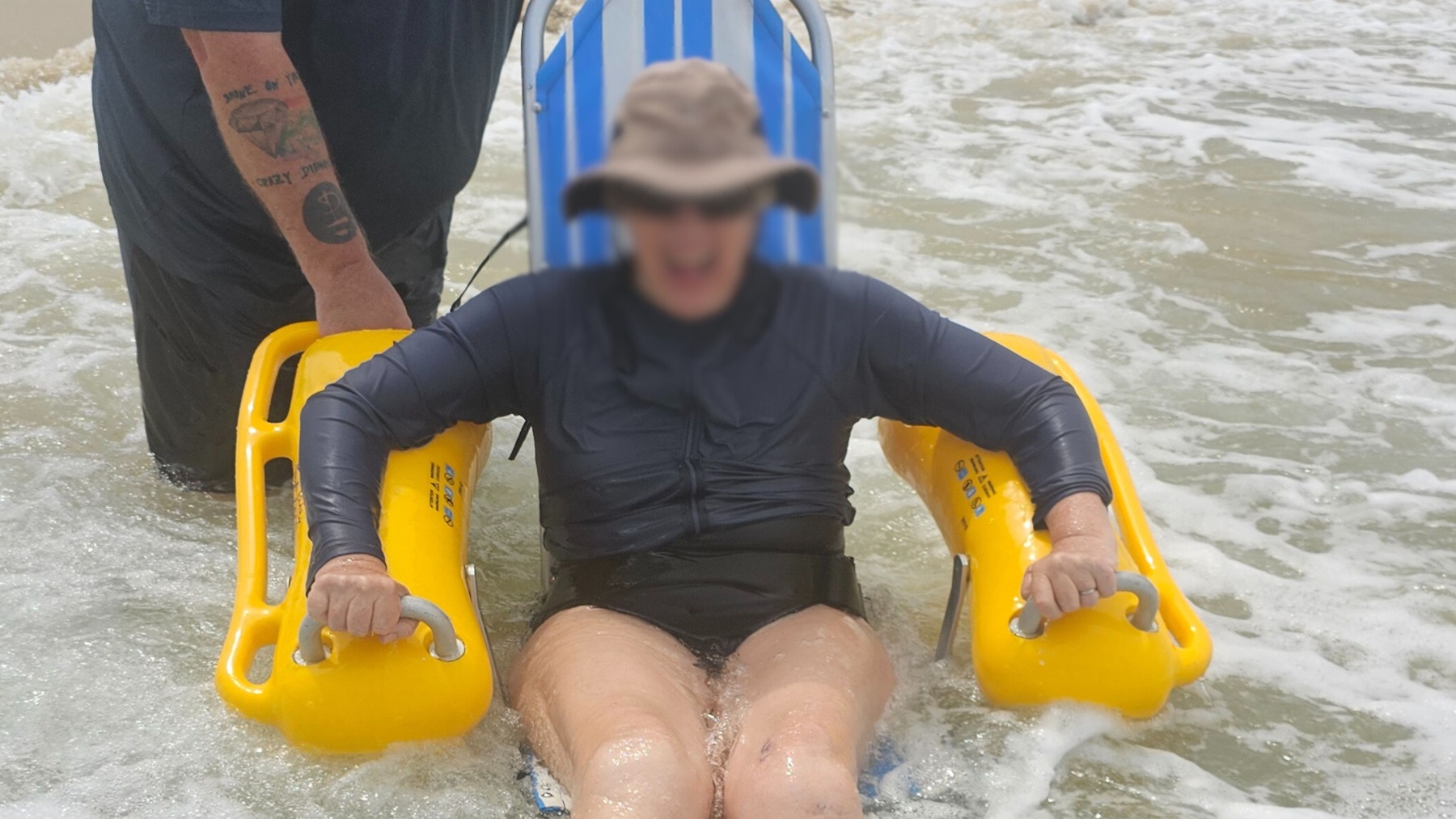 A person wearing a grey hat and dark long-sleeved clothing sits on a blue-and-white striped seat with yellow armrests, partially submerged in shallow water. Another person, only partially visible on the left, is standing behind and holding the chair. The foreground is covered with white foamy water from the sea, with sandy textures faintly visible underneath. The background contains more shallow water extending outwards.