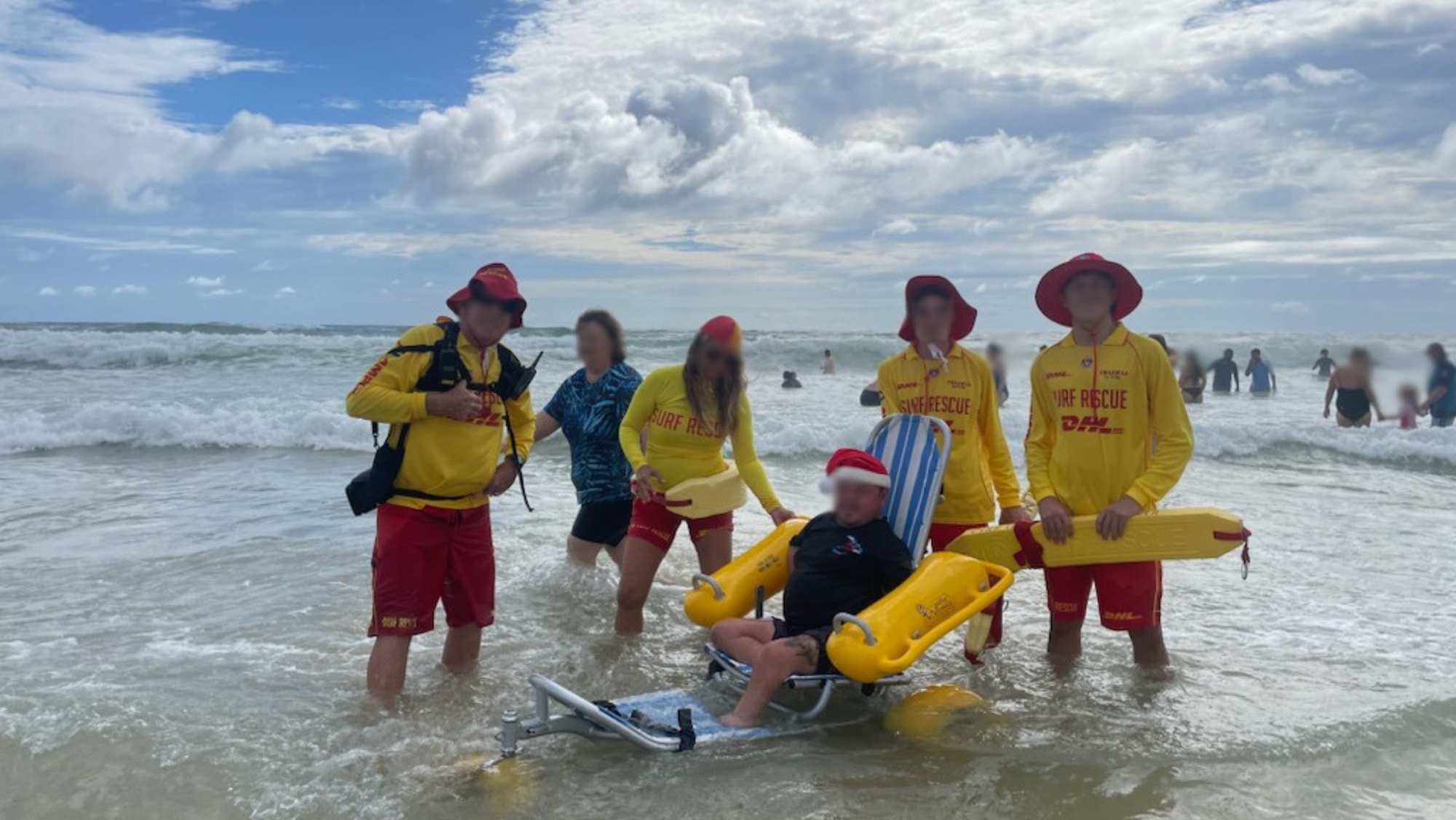 Five people stand in shallow water with foamy waves in the background. Four of them wear yellow surf rescue shirts and red hats, standing to the left and right of a person seated in a blue-and-white striped beach wheelchair with yellow floats. The seated person is wearing a Santa hat and dark clothing. The foreground is covered with rippling water, and several people are visible further back in the sea.