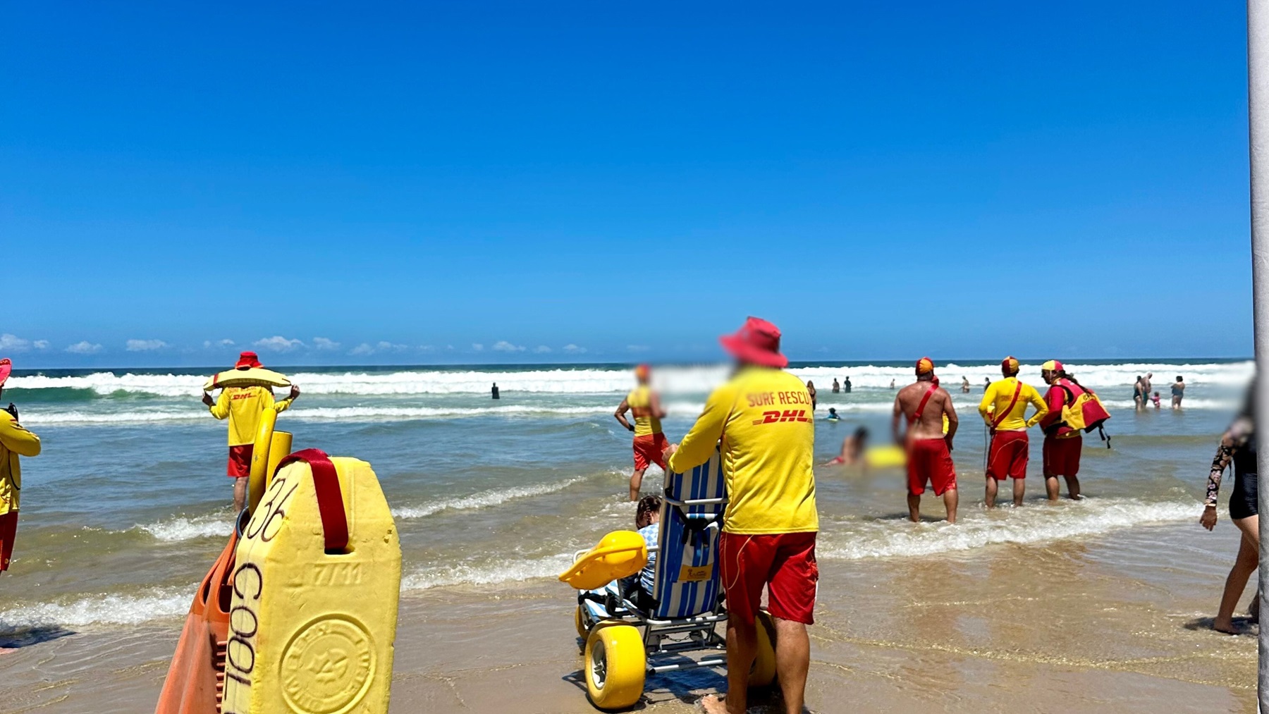 On the left, a yellow rescue board with attached straps leans upright in the sand. On the right, a man in a yellow and red surf rescue uniform stands next to a beach wheelchair with yellow wheels and blue-and-white striped seating. In the middle, several people in yellow and red uniforms stand in shallow water facing the waves, with others further out in the sea. The foreground is wet sand with shallow water, and waves are visible in the background.