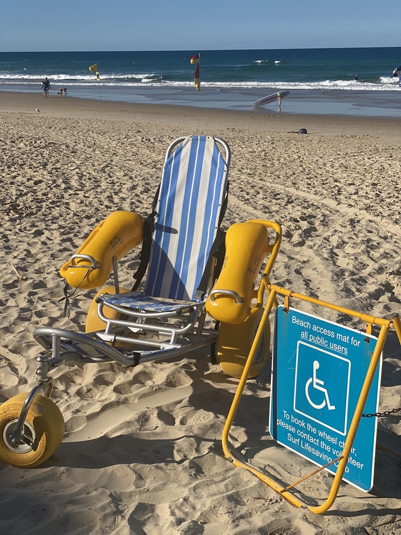 A blue-and-white striped beach wheelchair with yellow inflatable armrests and wheels is positioned on sandy ground in the foreground. To its right, a bright yellow-framed sign displays a blue accessibility notice with booking instructions. The background includes a lifeguard flag on the left, rippling waves, and two figures walking along the shoreline. The sand in the foreground is uneven with visible footprints.