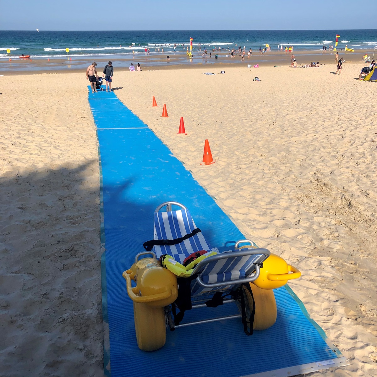 A yellow beach wheelchair with blue-and-white striped seating is in the foreground on a blue mat that stretches down the sandy beach toward the sea. On the left and right of the mat, the sand is smooth with scattered footprints. Orange cones line the right side of the mat. In the background, several people are in shallow water, with more swimmers further out. The floor is sandy with a textured blue mat, and the scene has no visible ceiling or overhead structures.