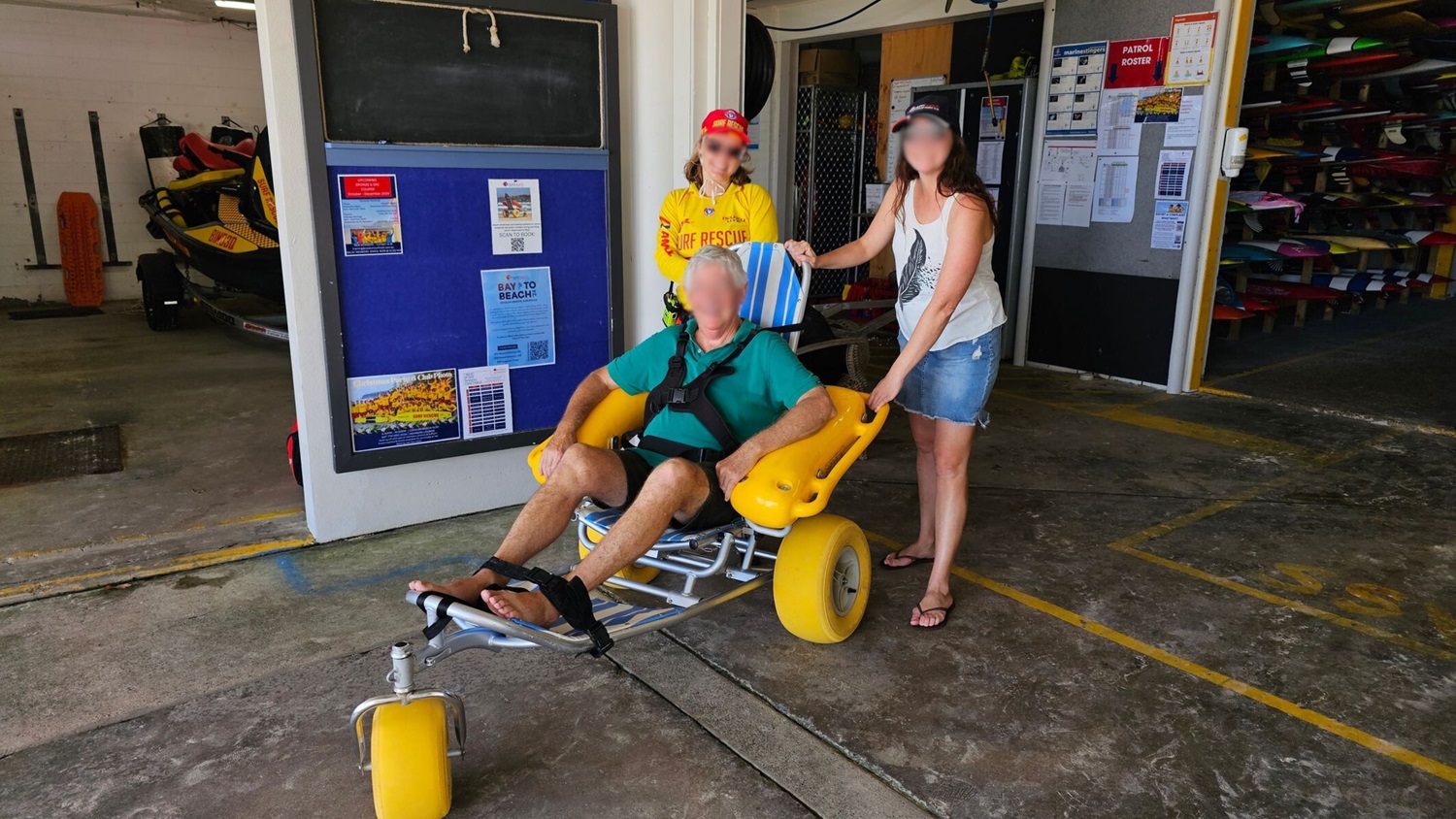 A man is seated in a beach wheelchair with large yellow wheels, wearing a green shirt and black shorts. A woman in a yellow lifeguard shirt and red cap stands to the left, while another woman in a white tank top and denim shorts stands to the right, holding the wheelchair. Behind them is a building interior with a blue notice board and various equipment. The floor is concrete with some painted lines.