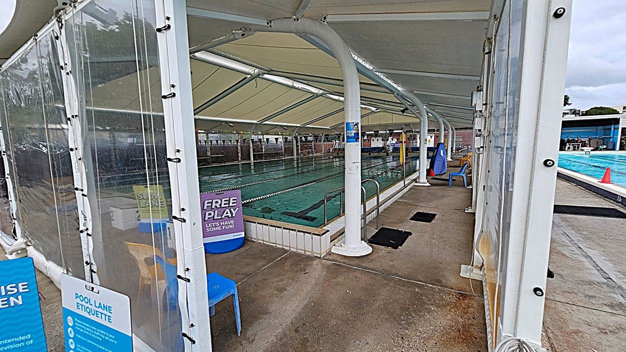 A swimming pool area with a covered structure on the left, featuring transparent walls and a white roof. A person in a yellow and red uniform stands near the entrance, where signs about pool etiquette and child supervision are displayed. The pool is on the right, with lanes marked and a few swimmers visible. The ground is a mix of concrete and green artificial turf. In the background, there are trees and a few buildings.