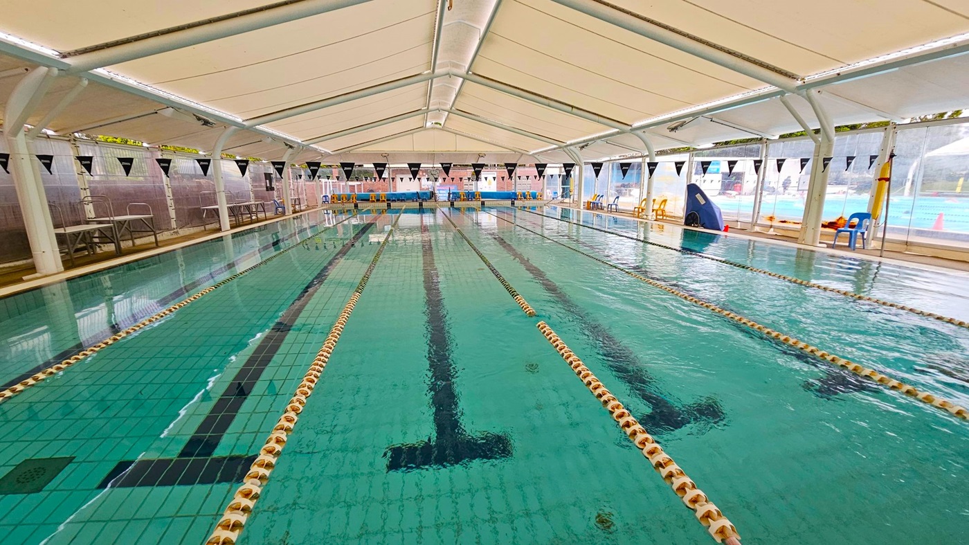 An indoor swimming pool with multiple lanes, separated by floating lane dividers. The pool is covered by a white tent-like structure with a high ceiling. On the left and right sides, there are transparent walls with black triangular flags hanging above. Several plastic chairs in various colours are placed along the sides. The floor of the pool is tiled in green with black lane markings.