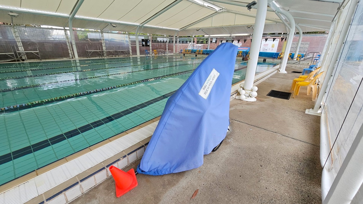 An indoor swimming pool with multiple lanes is on the left, covered by a white canopy. On the right, a blue cover labelled "PAL Portable Pool Lift" leans against a white column. Several yellow plastic chairs are lined up along the poolside, and a red traffic cone is placed near the pool's edge. The floor is concrete, and there are transparent panels on the right side.