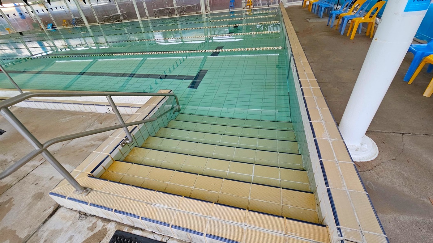 Steps leading into a swimming pool with greenish tiles. A metal handrail is on the left. The pool has lane dividers and is indoors, with a row of blue and yellow plastic chairs on the right side. The floor around the pool is concrete with some discolouration. A white column is visible near the chairs.