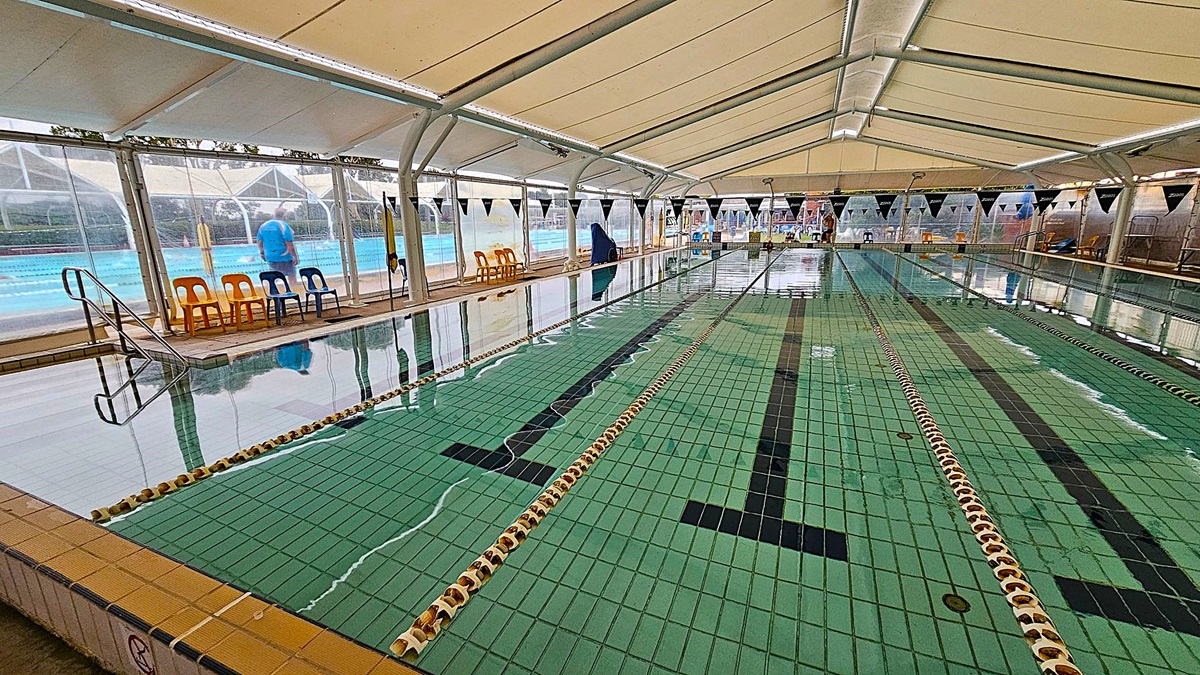 An indoor swimming pool with green-tiled lanes and black lane markings. The pool is covered by a large white tent structure with transparent side panels. On the left, there are several yellow and blue plastic chairs along the poolside. A metal ladder descends into the water. In the background, there are more chairs and a person walking outside the transparent panels. The ceiling is supported by white beams.