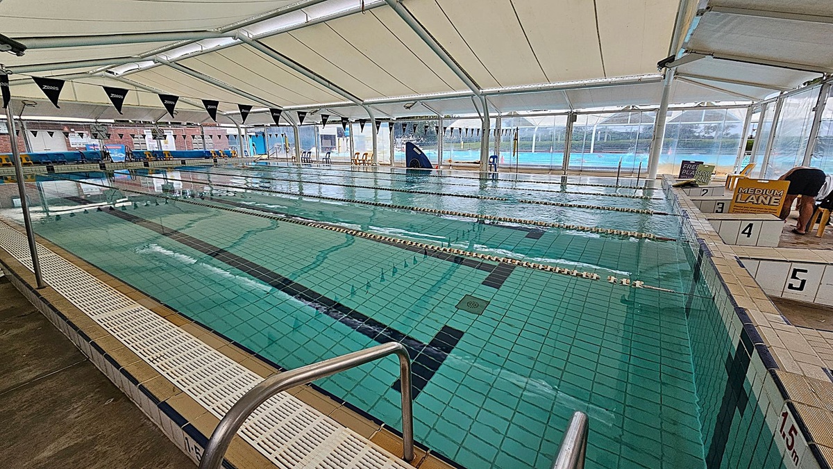 Indoor swimming pool with multiple lanes, each separated by lane ropes. The pool has a tiled floor with marked lanes. On the right, there are starting blocks numbered 2 to 5, with signs indicating "Free Play Slow Lane" and "Medium Lane". The ceiling is a white tent-like structure, and the walls are transparent, allowing a view of an outdoor pool area. There are chairs along the far side of the pool. A metal handrail is visible in the foreground.