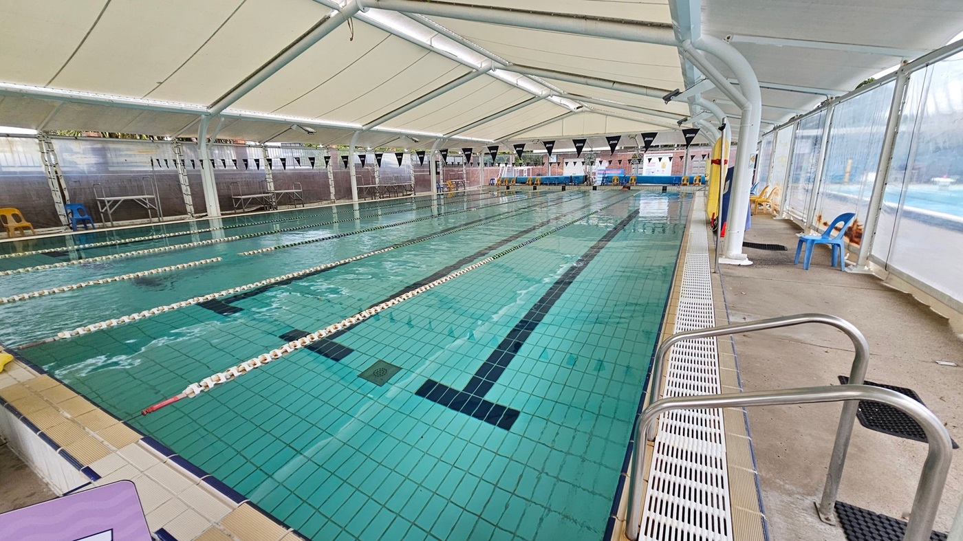 An indoor swimming pool with multiple lanes, separated by floating lane dividers. The pool is covered by a large, translucent canopy. On the right, there are metal handrails leading into the water and a concrete walkway with blue plastic chairs. The left side features a tiled pool edge and a few more chairs. The background shows a wall with windows and some equipment. The ceiling is supported by white beams.