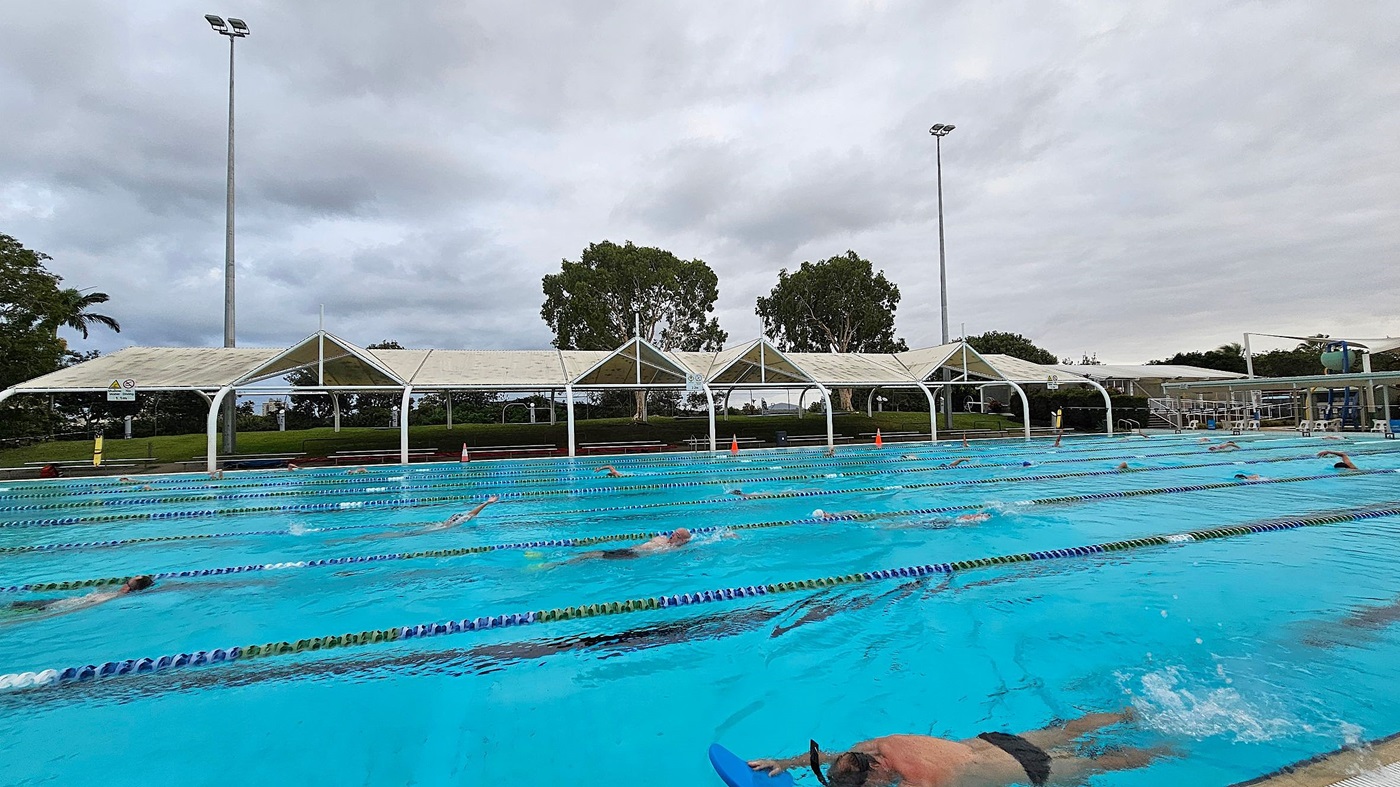An outdoor swimming pool with multiple lanes, where several swimmers are practising. On the right, a poolside area with a metal handrail and orange cones. In the background, there are white tents and a few buildings partially obscured by trees. A crane is visible behind the trees, indicating nearby construction. The pool's edge is tiled, and the water is clear and blue.