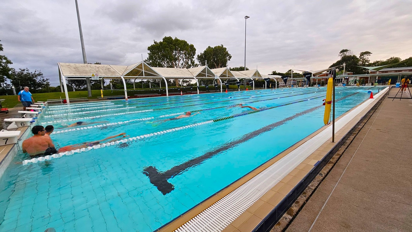 An outdoor swimming pool with several lanes, where people are swimming and sitting at the edge. On the left, a person in a blue shirt stands near the pool. The pool is surrounded by a concrete deck, and there are white shade structures with greenery in the background. A yellow rescue float is attached to a pole on the right side. The sky is overcast, and there are trees visible in the distance.