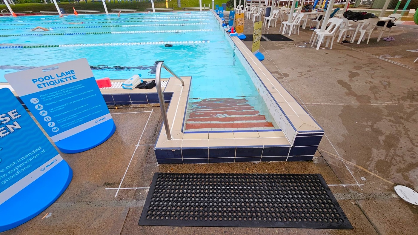 A swimming pool with lanes marked by floating dividers. In the foreground, there are steps leading into the pool with a metal handrail. A black rubber mat is on the wet concrete floor. To the left, a blue sign displays pool lane etiquette. On the right, several white plastic chairs are arranged along the poolside, with some personal items on them. The background shows more pool lanes and a swimmer in the water.