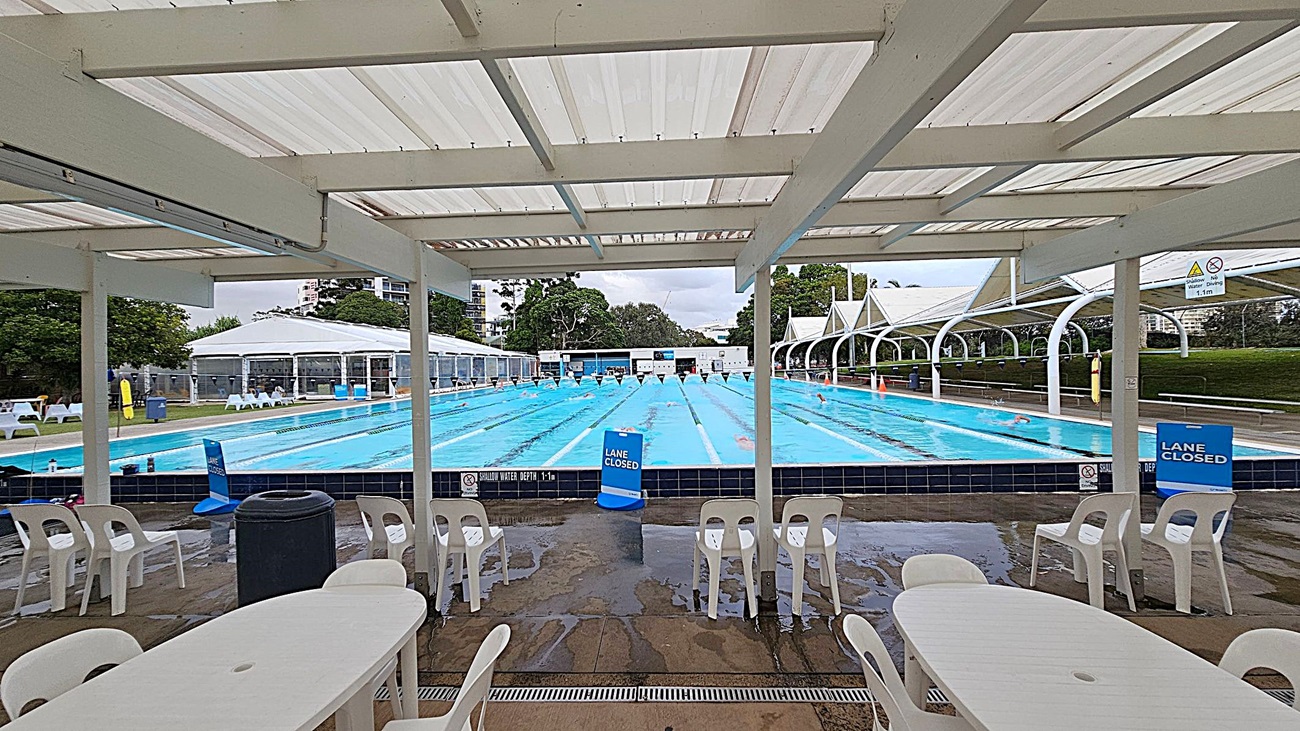 A covered outdoor swimming pool area with a long pool in the centre, divided into lanes with floating markers. Signs indicate some lanes are closed. White plastic tables and chairs are in the foreground on a wet concrete floor. The ceiling is a translucent material supported by beams. In the background, there are trees and a few buildings, with additional seating and structures on either side of the pool.