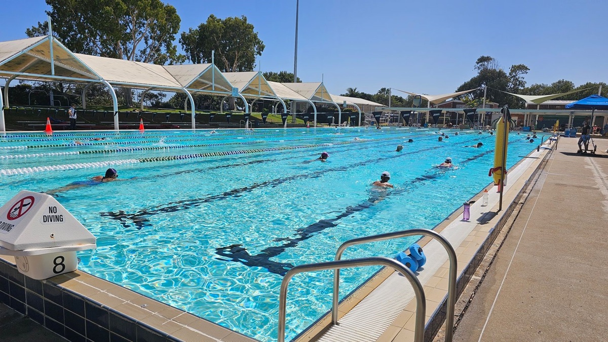 An outdoor swimming pool with multiple lanes, where several people are swimming. On the left, a "No Diving" sign is visible near lane 8. The pool is surrounded by a tiled deck, and on the right, there is a concrete walkway with a few items like a water bottle and a flotation device. In the background, there are shaded seating areas with white canopies and trees.