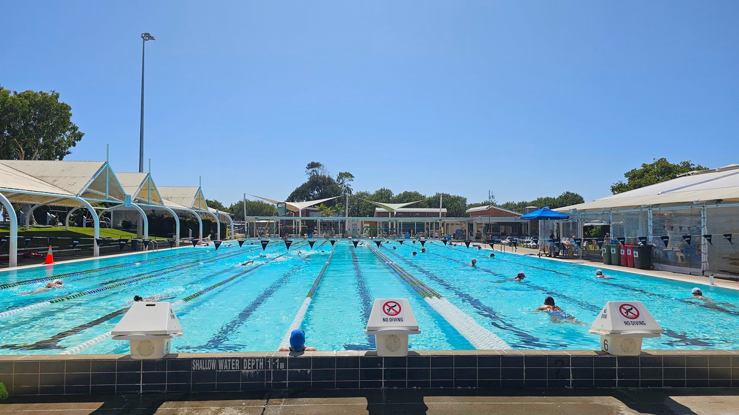 An outdoor swimming pool with multiple lanes, where several people are swimming. On the left, there are shaded seating areas with white canopies and a grassy section. On the right, there are more shaded areas, a blue umbrella, and bins. In the background, trees and a building are visible. The pool has signs indicating "No Diving" and a shallow water depth of 1.1 metres.