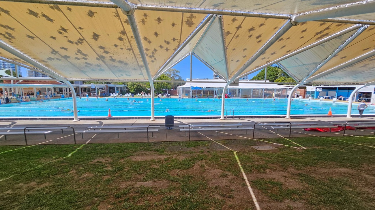 A large outdoor swimming pool with lanes is visible in the centre, surrounded by a paved area with benches. The pool is covered by a canopy with a pattern of dark shapes. In the background, there are buildings and trees. The foreground shows a grassy area with patches of dirt. Swimmers are in the pool, and a few people are walking around the perimeter.