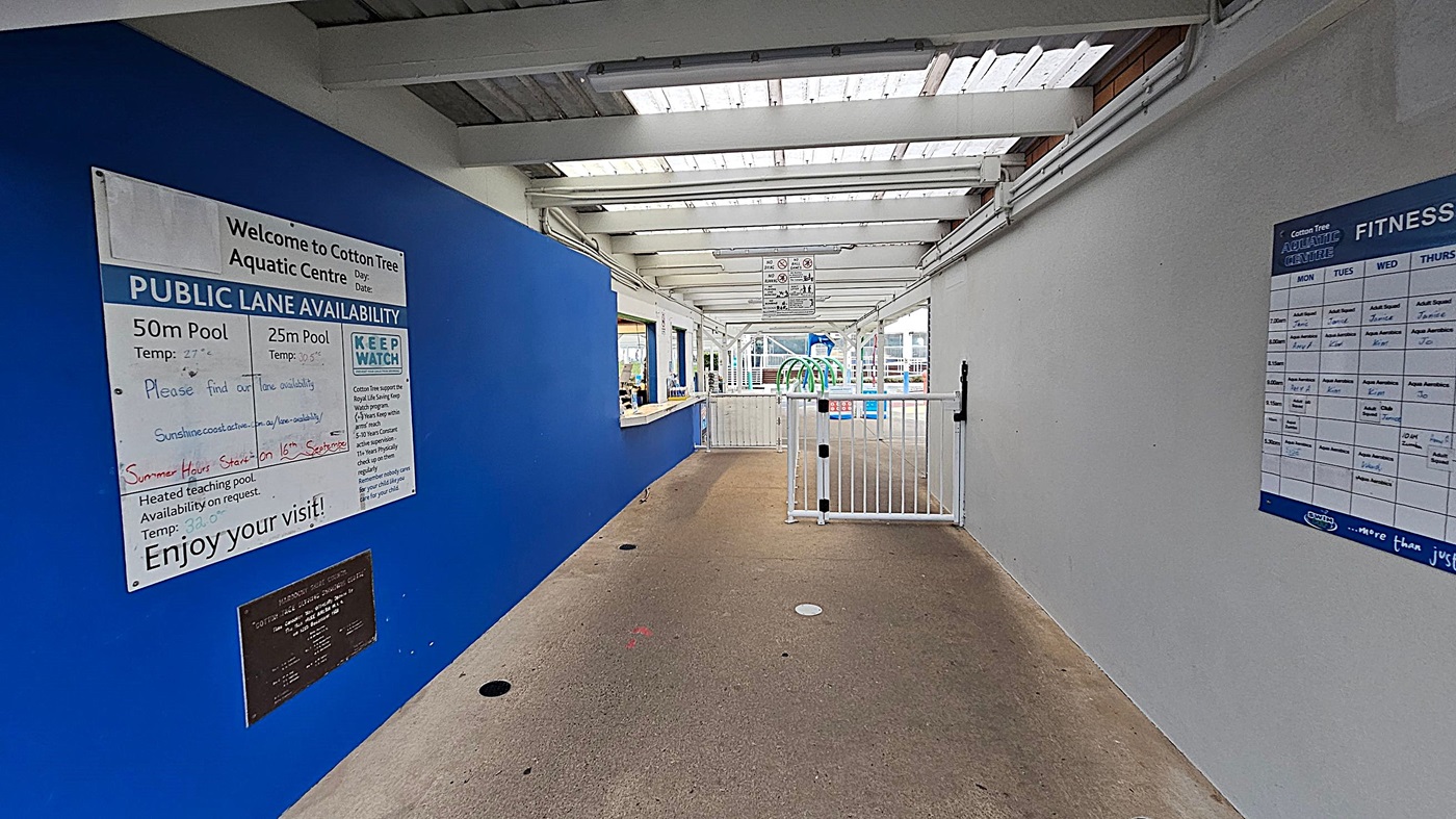 Entrance to the Cotton Tree Aquatic Centre with a sign above the doorway. Two bicycles are parked on the left against the wall, near a board displaying opening hours. A white bench and a shopping trolley are on the right. The building has a flat roof with a row of small circular vents. The ground is a mix of concrete and grass patches. A "No Smoking" sign is visible on the wall.