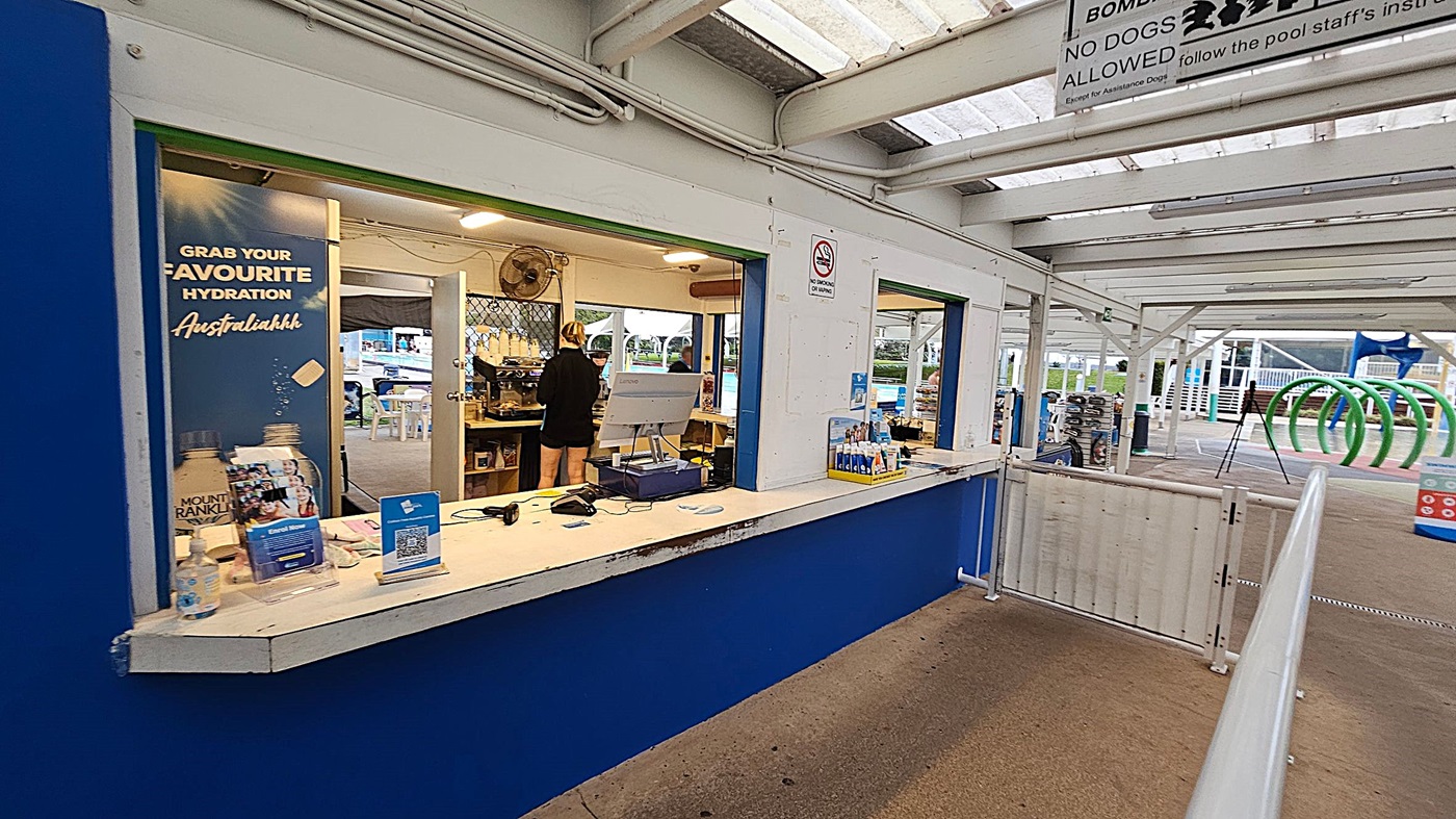 A kiosk with a blue and white counter on the left, displaying a sign promoting hydration. Inside, a person is working near a cash register and a coffee machine. The ceiling is covered with translucent panels. On the right, a fenced-off area leads to an outdoor space with green circular structures and a sign prohibiting dogs. The floor is a mix of concrete and textured surfaces.