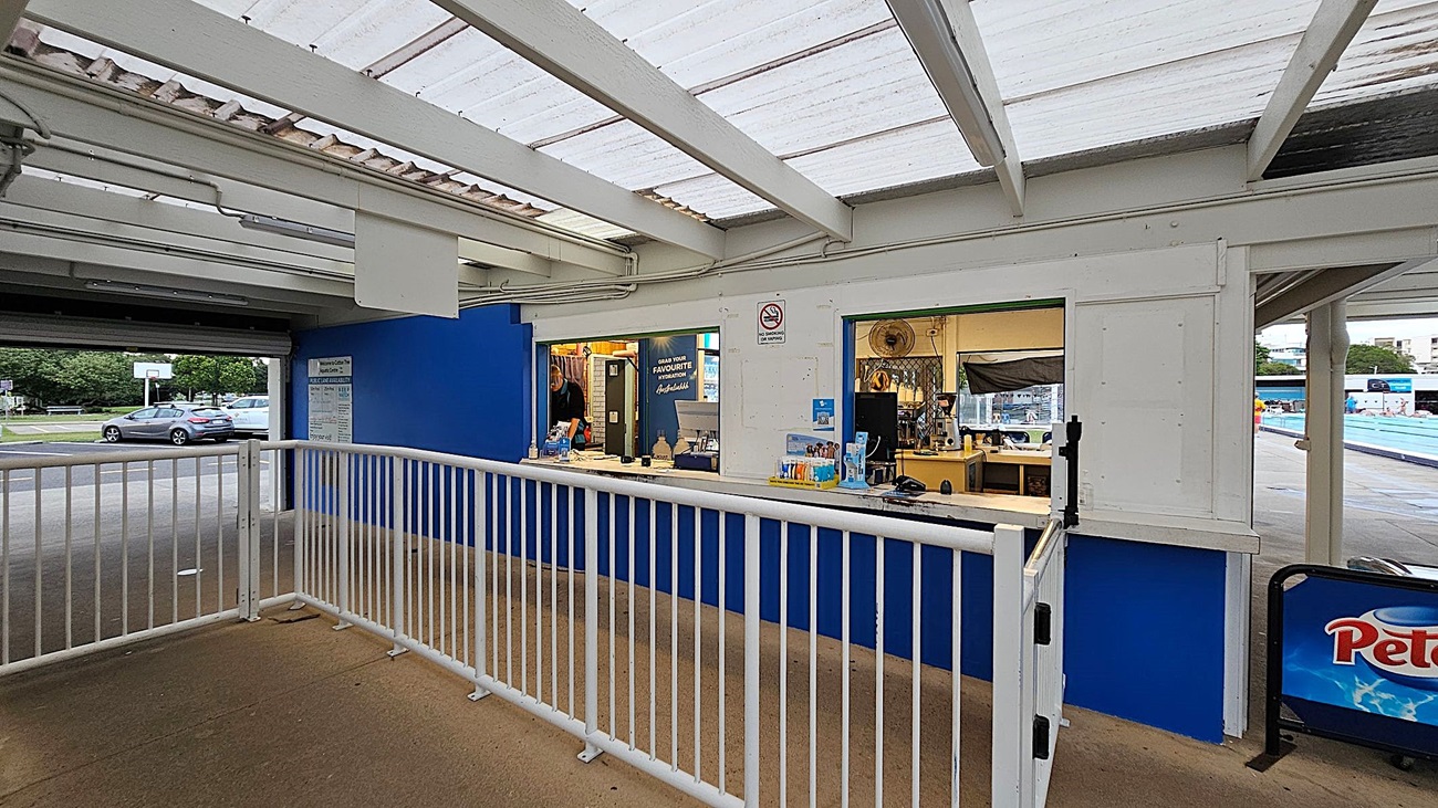 A kiosk with a blue and white exterior, featuring a service window with various items displayed. A white railing runs in front of the kiosk. To the right, there's a glimpse of a swimming pool area with people. The ceiling is made of translucent panels, allowing light through. On the left, a car is parked near a basketball hoop, with trees in the background. A signboard with "Peters" is visible on the right side.